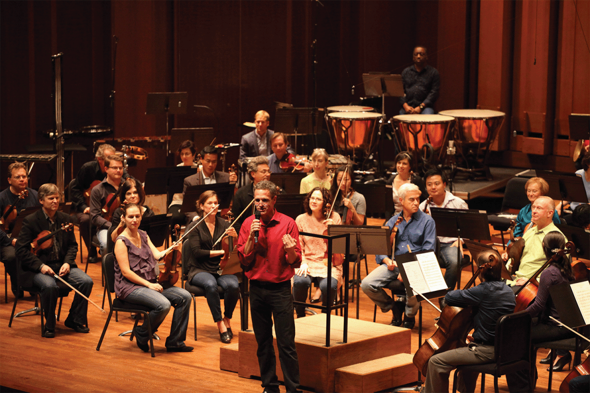 conductor speaking on microphone with the orchestra looking at him speak